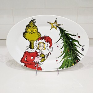 May include: Oval white ceramic platter with a colourful illustration of the Grinch holding a small girl, with a Christmas tree and star. The Grinch is green with a red suit, and the girl wears a red hat and pink dress. Ornaments decorate the tree.