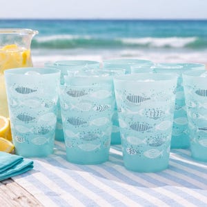 May include: A set of light blue drinking glasses with a fish pattern, arranged on a striped tablecloth. A glass pitcher of lemonade and fresh lemons are also visible, with the ocean in the background. A refreshing summer theme.