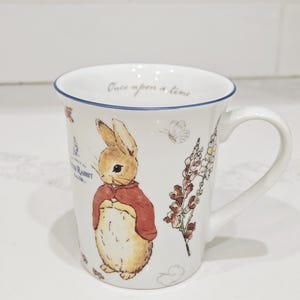 May include: White ceramic mug featuring a Peter Rabbit illustration with a red jacket and floral accents. The mug has a blue rim and the words "Once upon a time" are printed on the inside rim.