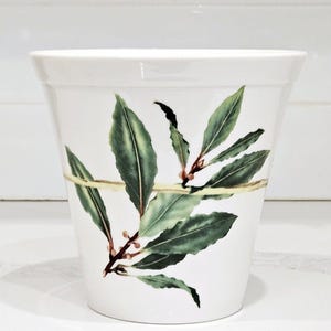 May include: White ceramic planter with a green and brown bay leaf design. The planter has a wide mouth and a rounded bottom.