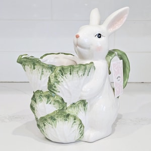 May include: A white ceramic bunny-shaped planter with a green and white cabbage design. The bunny has pink cheeks and a small tag attached. This decorative planter is ideal for Easter or spring decor.