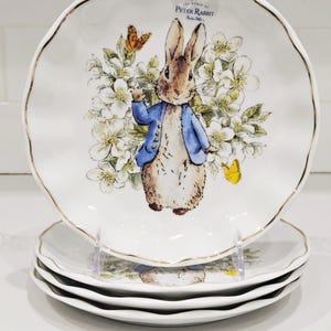 Puede incluir: Una pila de cuatro platos de cerámica blanca con borde festoneado y ribete dorado. Cada plato presenta una ilustración de acuarela de Peter Rabbit con una chaqueta azul, rodeado de flores y mariposas. Los platos son de "The World of Peter Rabbit."
