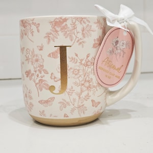Pode incluir: Caneca de cerâmica creme com um padrão floral rosa e a letra dourada "J". A caneca tem uma base castanha clara e uma alça branca. Uma etiqueta rosa com a palavra "Artisanal" é presa com uma fita branca.