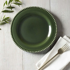 May include: A dark green ceramic plate with a beaded edge. A white napkin and a silver fork are on the right side of the plate. A sprig of green leaves is in the upper left corner. The plate is on a white wooden surface.