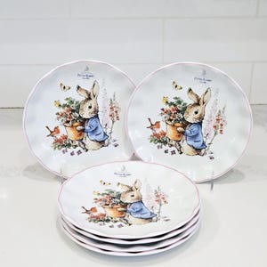 May include: Set of white ceramic plates with a scalloped edge and a pink trim. Each plate features a Peter Rabbit illustration with flowers and butterflies. The plates are stacked and arranged on a white surface.