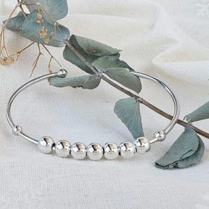 Op de afbeelding: Een zilveren armband met acht glanzende, ronde kralen. Het open ontwerp wordt getoond op een witte, getextureerde ondergrond met groene bladeren. Een eenvoudig, elegant sieraad.