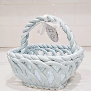 Puede incluir: Una cesta de cerámica azul claro con un diseño tejido y un asa trenzada. Una cinta blanca y una etiqueta que dice "Simply Spring" están unidas al asa. La cesta está sobre una superficie blanca.