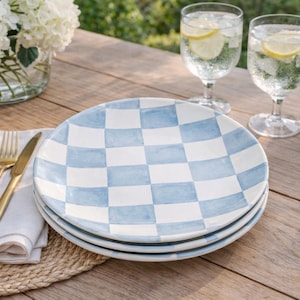 Puede incluir: Tres platos de cerámica con un diseño de cuadros azules y blancos. Los platos están sobre una mesa de madera, junto con cubiertos dorados, una servilleta blanca y vasos de agua con rodajas de limón. También hay una cesta de pan y un cuenco de arándanos.