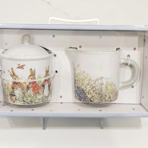 May include: A ceramic sugar bowl with a lid and a creamer jug set. The sugar bowl and jug feature a whimsical illustration of rabbits in a field. The set is presented in a light blue box with a white interior.