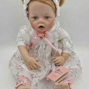 May include: A realistic baby doll with blonde pigtails, dressed in a white dress with pink rose embroidery and a matching headband. The doll has blue eyes and a pink bow. A tag from Paradise Galleries is visible.