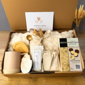 May include: A gift box filled with a variety of items, including a mug, chocolates, a spoon, and a decorative card. The box contains a white blanket, a wooden scoop, and a small bag. The chocolates are labeled "Macadamias" and weigh 100g.
