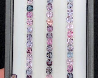 May include: A collection of small, faceted, cushion-cut gemstones arranged in three vertical rows and two small clusters within a gray display tray. The stones feature a variety of translucent colors including shades of pink, purple, blue, and gray.