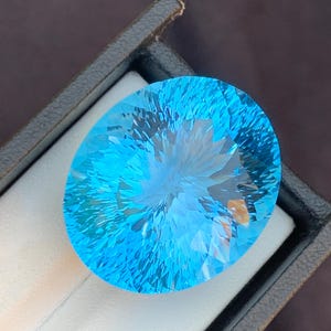 Op de afbeelding: Een grote, ovaal geslepen, briljant blauwe edelsteen. De edelsteen is gefacetteerd om licht te reflecteren, waardoor een steruitbarsting-effect ontstaat. Het is geplaatst in een zwarte sieradendoos met een wit interieur, tegen een donkere achtergrond.