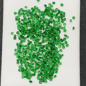 May include: A collection of small, round, vibrant green gemstones scattered on a white surface. The gems are contained within a small, open, brown container, suggesting a presentation or storage setting. The overall aesthetic is one of natural beauty and preciousness.