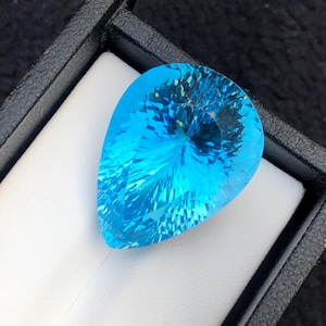 Peut inclure: Une grosse pierre précieuse facettée en forme de poire, d'un bleu clair éclatant. La gemme est présentée dans un écrin noir avec un intérieur blanc, créant un contraste saisissant. Les facettes captent la lumière, révélant la brillance et la clarté de la gemme.