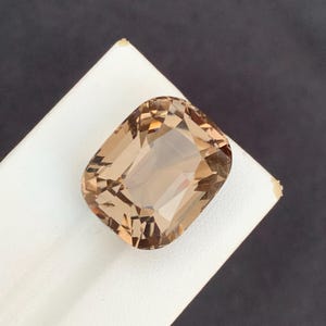 May include: A faceted, rectangular gemstone with a warm, light brown hue. The gem is set against a white surface, with a dark gray background. The cut of the stone reveals multiple facets.