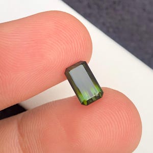 May include: A rectangular-cut, green gemstone with a dark, reflective interior. The gem is held between two fingers, showcasing its polished facets and colour variations. The gemstone's shape is a classic emerald cut, ideal for jewellery.