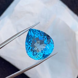 May include: A faceted, pear-shaped, bright blue gemstone is held by tweezers against a white background. The gem has a complex internal structure, reflecting light. The tweezers are silver-colored.
