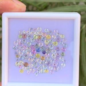 Pode incluir: Uma coleção de pequenas pedras preciosas redondas em uma variedade de cores, incluindo rosa, amarelo, azul e verde, é exibida em uma moldura quadrada branca. As pedras preciosas são colocadas sobre um fundo lilás claro, criando uma exibição colorida e atraente.