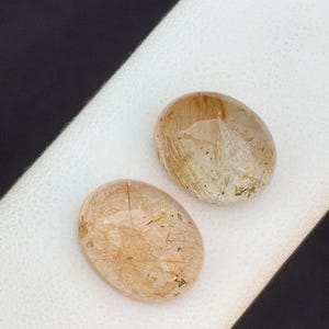 以下が含まれることがあります： 金茶色の色合いと内部インクルージョンを持つ2つの楕円形の宝石。石は黒い背景の白い表面に置かれています。宝石は研磨され、半透明の外観をしています。