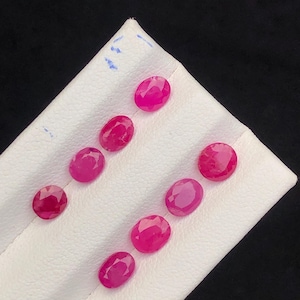 May include: Nine oval-cut, vibrant red gemstones are arranged on a white, textured surface against a black background. The gemstones exhibit a polished, reflective surface, showcasing their clarity and rich color. These gemstones are likely rubies, perfect for jewelry making.