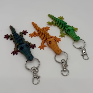 Pode incluir: Três chaveiros de jacaré coloridos. Um é verde, outro laranja e outro azul. Cada jacaré tem um chaveiro e um fecho prateados. Os chaveiros são feitos de um material texturizado.