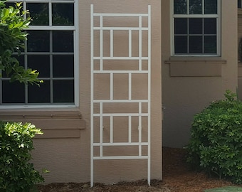 May include: White metal trellis with a geometric design, standing in front of a beige house with two windows. The trellis is about 6 feet tall and 3 feet wide.
