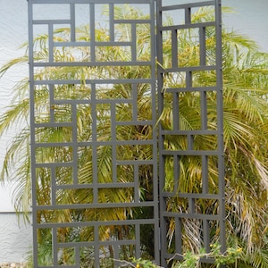 May include: Two sections of a dark gray metal garden trellis with a geometric design. The trellis is positioned in front of a backdrop of green palm fronds and small yellow flowers.