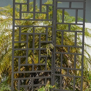May include: A black metal trellis with a geometric design, standing in front of a green bush.