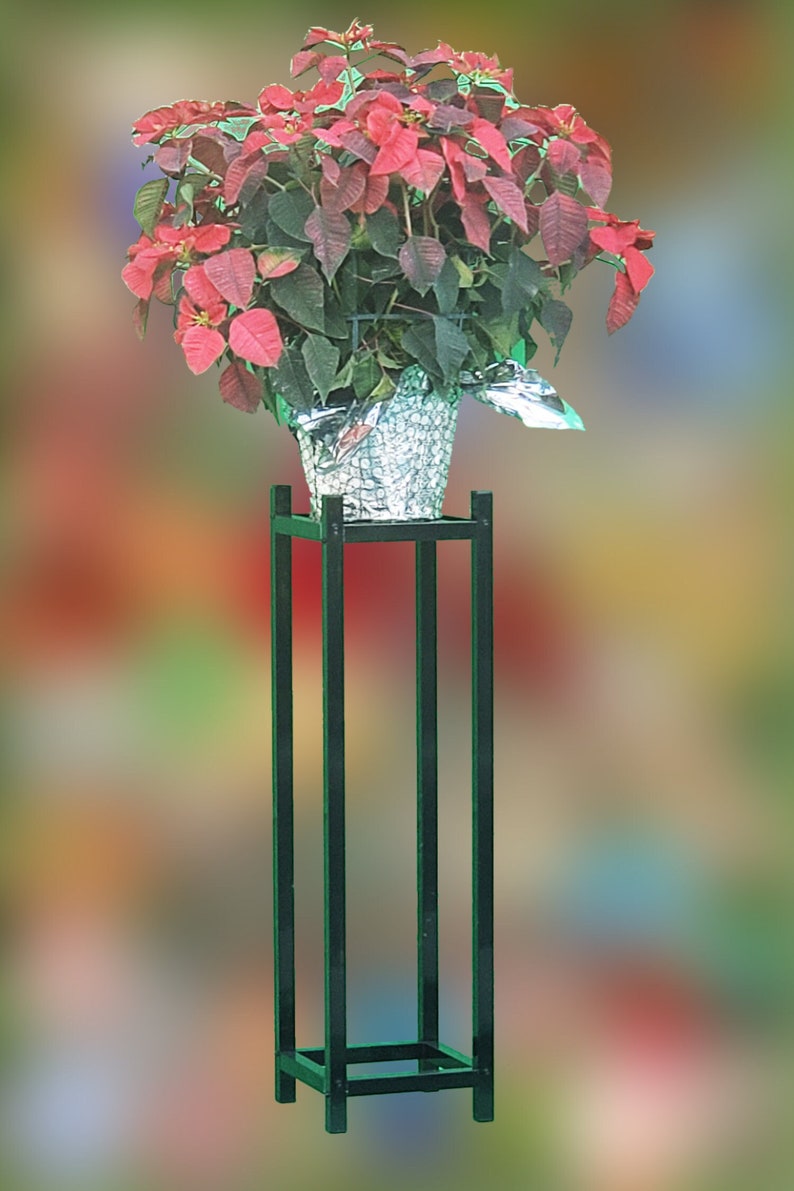 May include: A potted poinsettia plant with vibrant red bracts and dark green leaves sits on a tall, black metal plant stand. The plant is in a silver foil-wrapped pot, creating a striking contrast.
