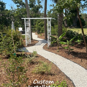 May include: A white, decorative garden archway with a geometric design stands at the entrance of a winding gravel path. Lush greenery and palm trees frame the path, with the text "Custom Installation" at the bottom.