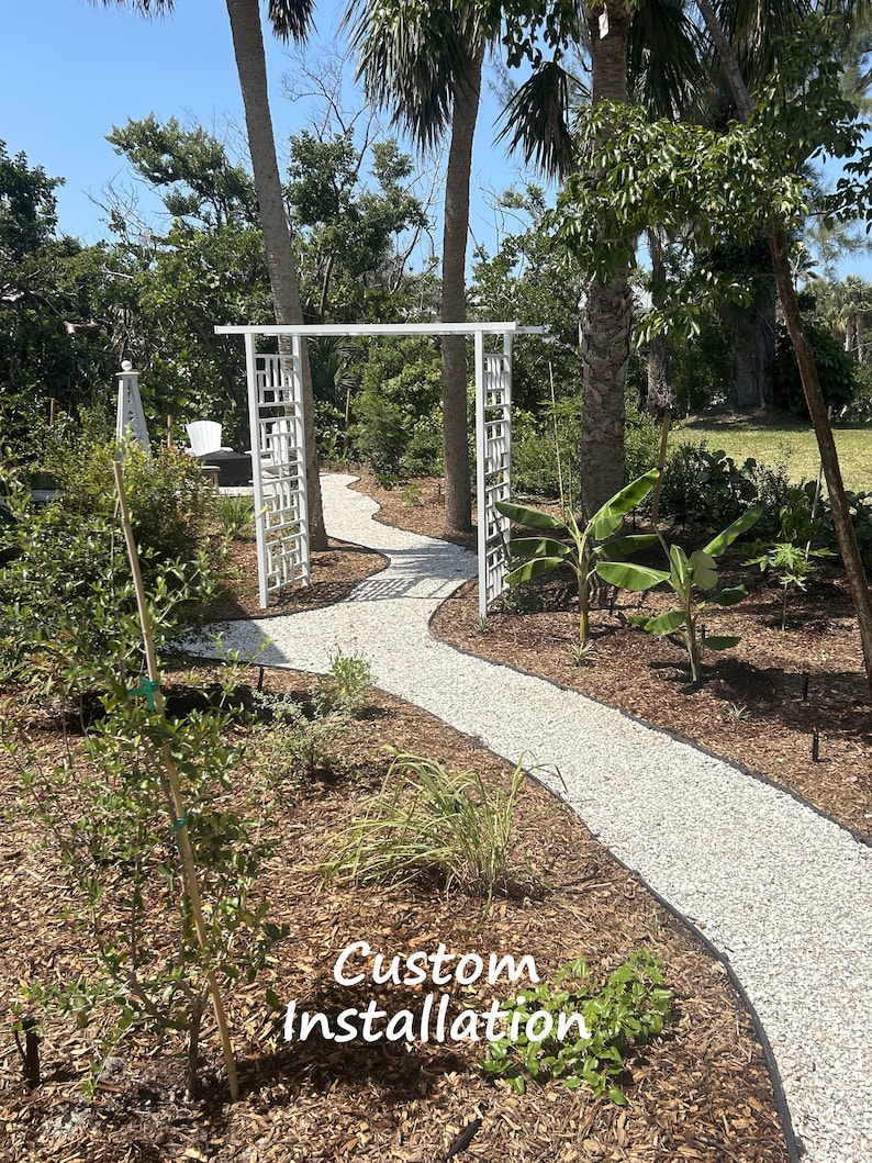 May include: A white, decorative garden archway with a geometric design stands at the entrance of a winding gravel path. Lush greenery and palm trees frame the path, with the text "Custom Installation" at the bottom.