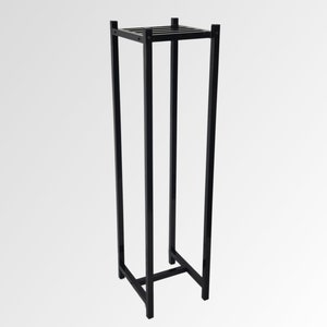 May include: A tall, black metal plant stand with a square top and base. The stand has a rectangular frame with vertical supports and a slatted top. The design is minimalist and modern, suitable for displaying plants or decorative items.