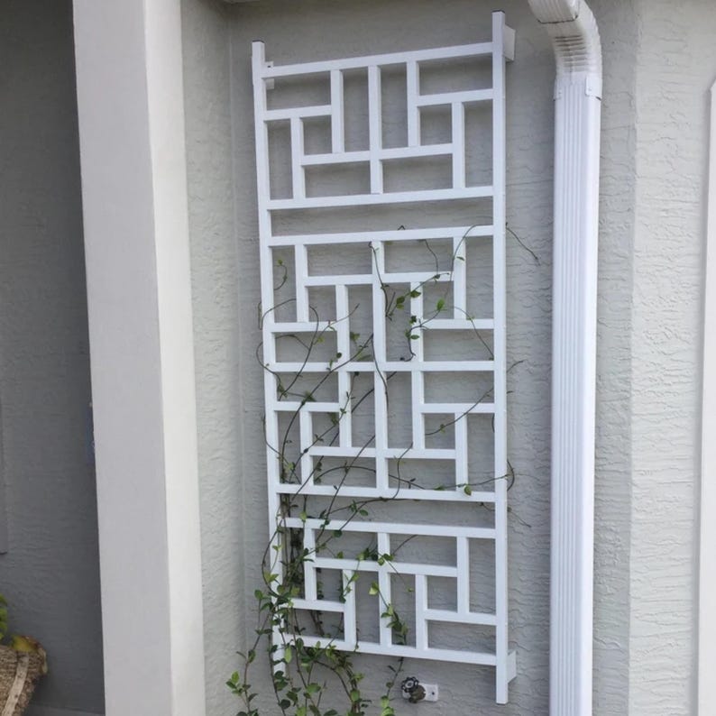 May include: A white, geometric trellis mounted on a light gray wall. The trellis has a rectangular shape with a repeating pattern of squares and rectangles. Green vines are growing through the trellis, adding a natural element to the design.