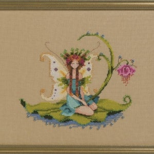 May include: A cross-stitch embroidery of a fairy with wings sitting on a leaf. The fairy has long brown hair, a blue dress, and a flower crown. The leaf is green and has a vine with a pink flower growing from it.