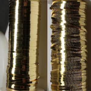 May include: Two spools of shiny, gold-colored ribbon. The ribbon is tightly wound on the spools, creating a stacked appearance. The ribbon reflects light, giving it a metallic sheen. The spools are set against a neutral background.