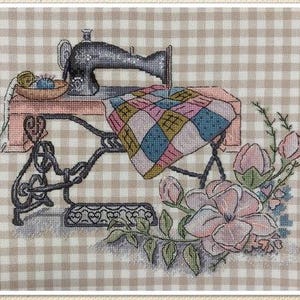 May include: Cross-stitch image of a vintage sewing machine with a colorful quilt draped over the table. A bowl of yarn and thread sits on the table. Pink flowers and green leaves are in the lower right corner. The background is a tan and white checkered pattern.