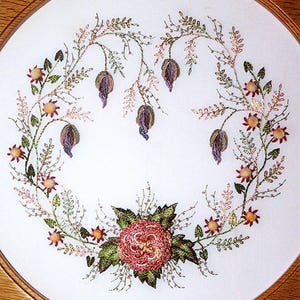 May include: A white embroidery hoop with a floral design. The design features a large pink flower in the center surrounded by smaller flowers and leaves in shades of green, pink, and purple.