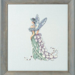 May include: A cross-stitch pattern of a fairy with blue wings and a green dress decorated with white flowers. The fairy is standing on a light blue background.