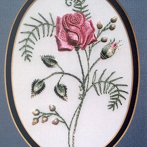 May include: A pink rose with green leaves and pearl-like beads embroidered on a white fabric background. The embroidery is framed in a black and gold oval frame.