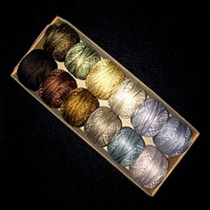 May include: A collection of 12 small balls of yarn in various colors and textures, including brown, gold, green, and gray. The balls of yarn are arranged in a row in a wooden box.
