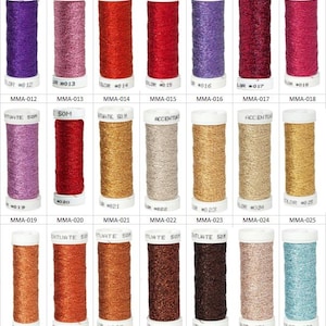 May include: A collection of twenty-one spools of metallic embroidery thread arranged in three rows. Each spool features a white plastic top and base with a label reading ACCENTUATE 50M and a unique color number. The threads display a shimmering finish.