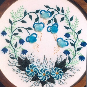 May include: Embroidered hoop art featuring a wreath of blue and green flowers. The flowers are made with a variety of stitches and have a 3D effect.