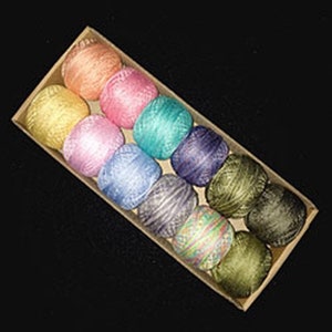 May include: A box of 12 colorful yarn balls in various shades of pink, blue, green, purple, and yellow. The yarn balls are arranged in a row in a cardboard box.