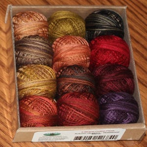 Puede incluir: Una caja con 12 carretes de hilo de algodón de 3 hebras en varios colores otoñales. Los colores incluyen tonos de marrón, naranja, rojo y verde. La caja tiene la inscripción "Autumn Outings - Rosewood."