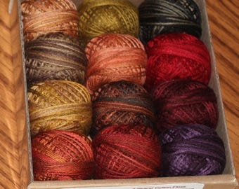 Könnte beinhalten: Eine Schachtel mit 12 Spulen 3-fädiger Baumwoll-Stickerei in verschiedenen Herbstfarben. Die Farben umfassen Brauntöne, Orange, Rot und Grün. Auf der Schachtel steht "Autumn Outings - Rosewood."