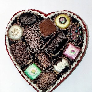May include: A heart-shaped box filled with embroidered chocolates. The chocolates are made of different colors and textures, including brown, green, pink, and white.