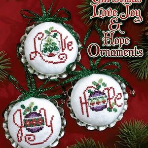 May include: Three Christmas ornaments with cross-stitch designs on bottle caps. The ornaments feature the words "Love", "Joy", and "Hope" with a stylized Christmas tree or ornament design.