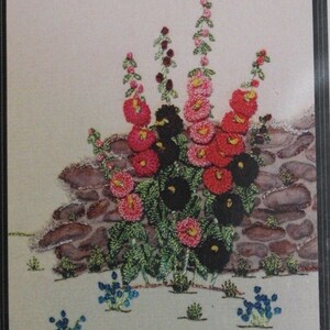May include: Embroidered wall art featuring a cluster of pink, red, and black flowers with green leaves, set against a brown stone wall. The flowers are arranged in a vase-like shape, with smaller blue flowers at the base. The design area is 5 inches by 5 inches.