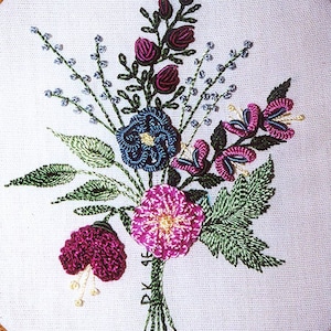 May include: A close-up of a hand-stitched embroidery hoop featuring a colorful floral bouquet with pink, purple, blue, and green flowers. The embroidery is done in a variety of stitches, including satin stitch, French knots, and stem stitch.